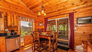A wooden cabin interior with a dining area, kitchen, and large sliding glass doors leading to a deck. - Shooting Star Ridge: Mountain View Cabin Near Cherokee with Hot Tub (Whittier)
