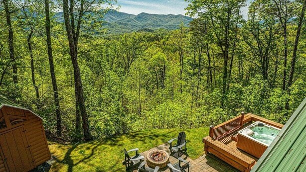 A wooden deck with a fire pit and chairs, surrounded by lush greenery and a mountainous backdrop. - Shooting Star Ridge: Mountain View Cabin Near Cherokee with Hot Tub (Whittier)