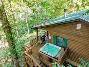 A wooden cabin with a hot tub on a deck surrounded by trees.