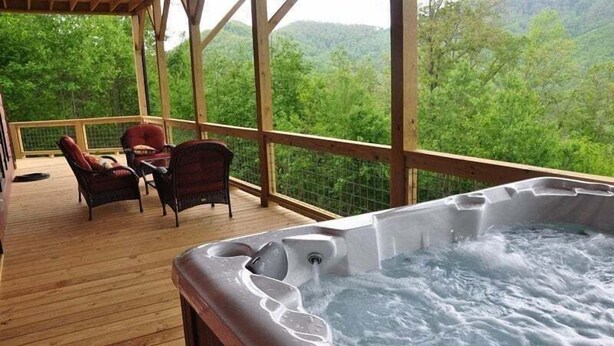 Un bain à remous sur une terrasse en bois, agrémentée de chaises et d’une table en osier, avec vue sur une zone boisée.