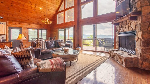 A spacious living room with a stone fireplace, leather furniture, large windows, and a view of the mountains. - A Walk in the Clouds (Bryson City)