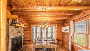 L’intérieur d’un chalet rustique en rondins de bois, avec une cheminée en pierre, une table à manger en bois et de grandes fenêtres offrant une vue sur l’extérieur.