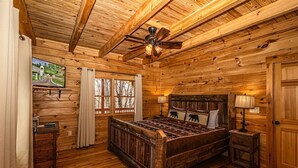 Une chambre à coucher dans une cabane en bois, avec un lit, un ventilateur de plafond, une table de chevet, une lampe et une fenêtre avec des rideaux.