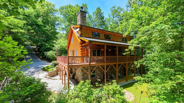 Une cabane en rondins dotée d’une terrasse et d’une cheminée en pierre, entourée d’une végétation luxuriante.