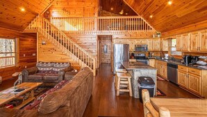 A spacious log cabin interior with a central staircase, a living area with a sofa and coffee table, and a kitchen with modern appliances.
