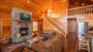 A log cabin interior with a stone fireplace, wooden staircase, and a kitchen area.