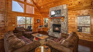 A cozy log cabin interior with a stone fireplace, wooden furniture, and large windows offering views of the outdoors.