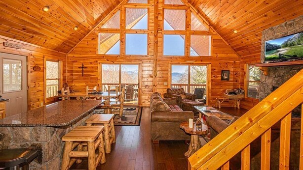 A log cabin interior with a high ceiling, wooden walls, a stone fireplace, a large TV, and a cozy seating area.