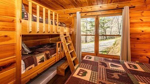 A cabin with wooden walls, a bunk bed, a ladder, and a large window with a view of trees.