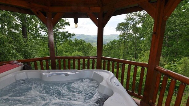 A hot tub with a wooden deck and railing, surrounded by lush greenery and mountains in the background. - Cherokee Ridge Retreat (Whittier)