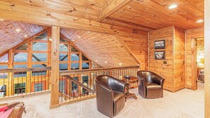 A wooden interior with a high ceiling, a balcony with glass railings, and two leather chairs.