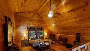 A cozy cabin bedroom with a wooden ceiling, a bed with patterned bedding, a nightstand, a chair, and a small table with a lamp. - 'Sunrise in the Smokies' - Where Mornings Begin Best (Bryson City)