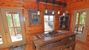 A wooden cabin interior with a wooden table, hanging light fixtures, a bear-themed wall decoration, and a bookshelf. - 'Sunrise in the Smokies' - Where Mornings Begin Best (Bryson City)