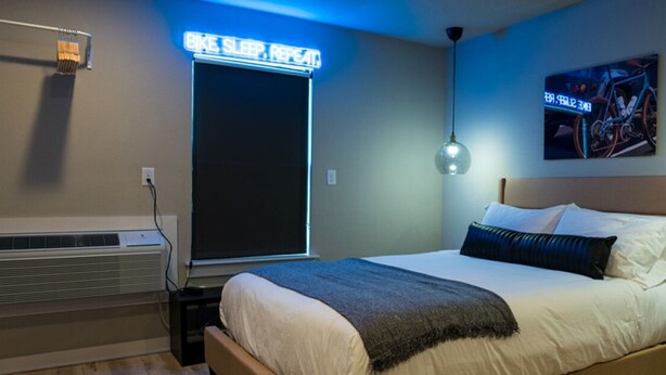 Un dormitorio con cama, aire acondicionado de pared, un letrero de neón y un cuadro con temática de bicicleta.
