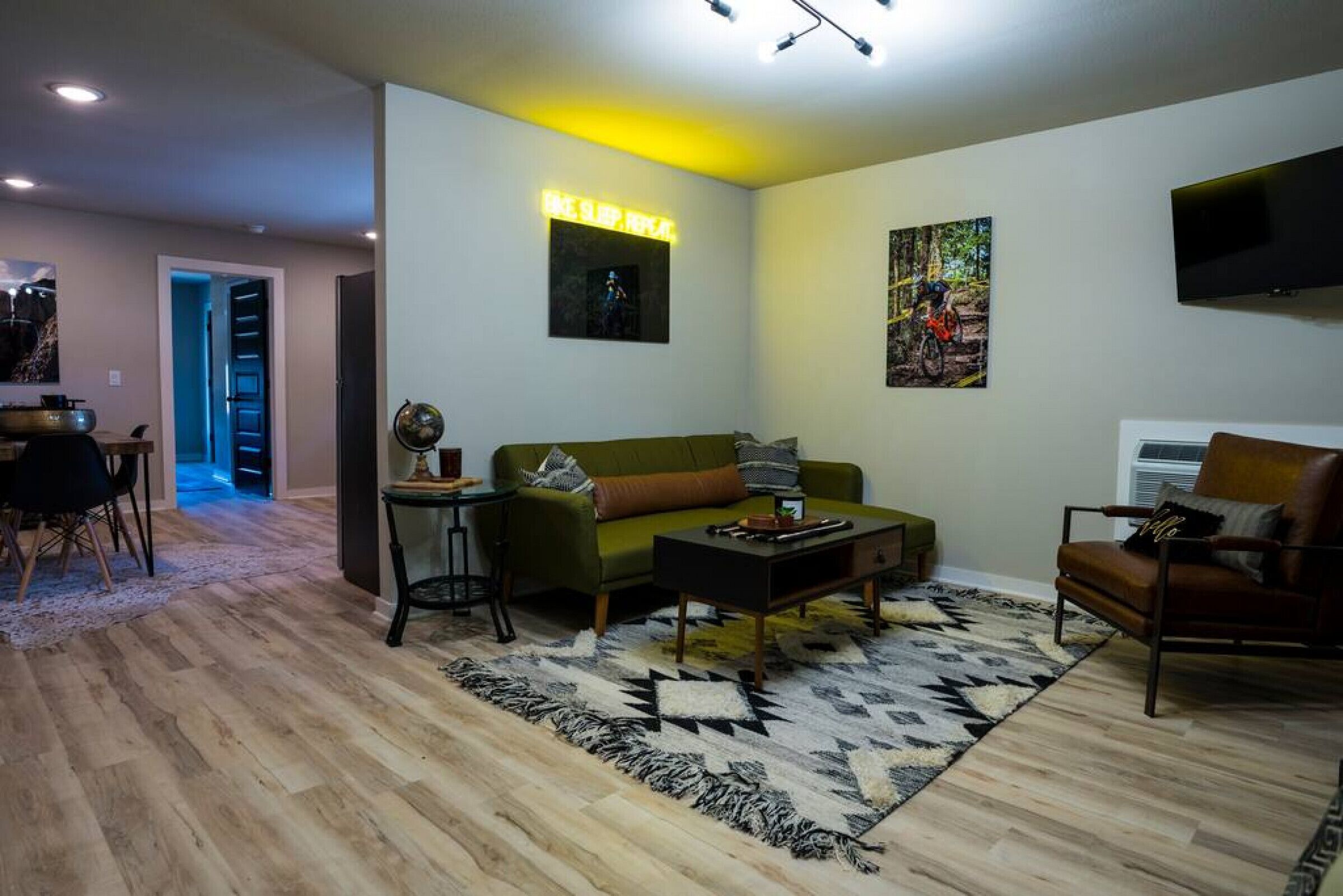 Uma sala de estar moderna com um sofá verde, uma mesa de centro e um tapete estampado. Há uma televisão fixada na parede e um quadro em cima.