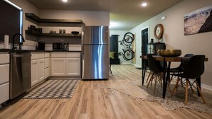 A modern kitchen with white cabinets, a stainless steel refrigerator, and a dining area with a wooden table and black chairs. - The Bike Inn #8, Boujee Bike Suite (Bentonville)