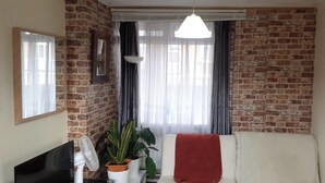 Un salon avec un mur en briques, un canapé blanc, un plaid rouge, un téléviseur, un ventilateur et des plantes en pot.
