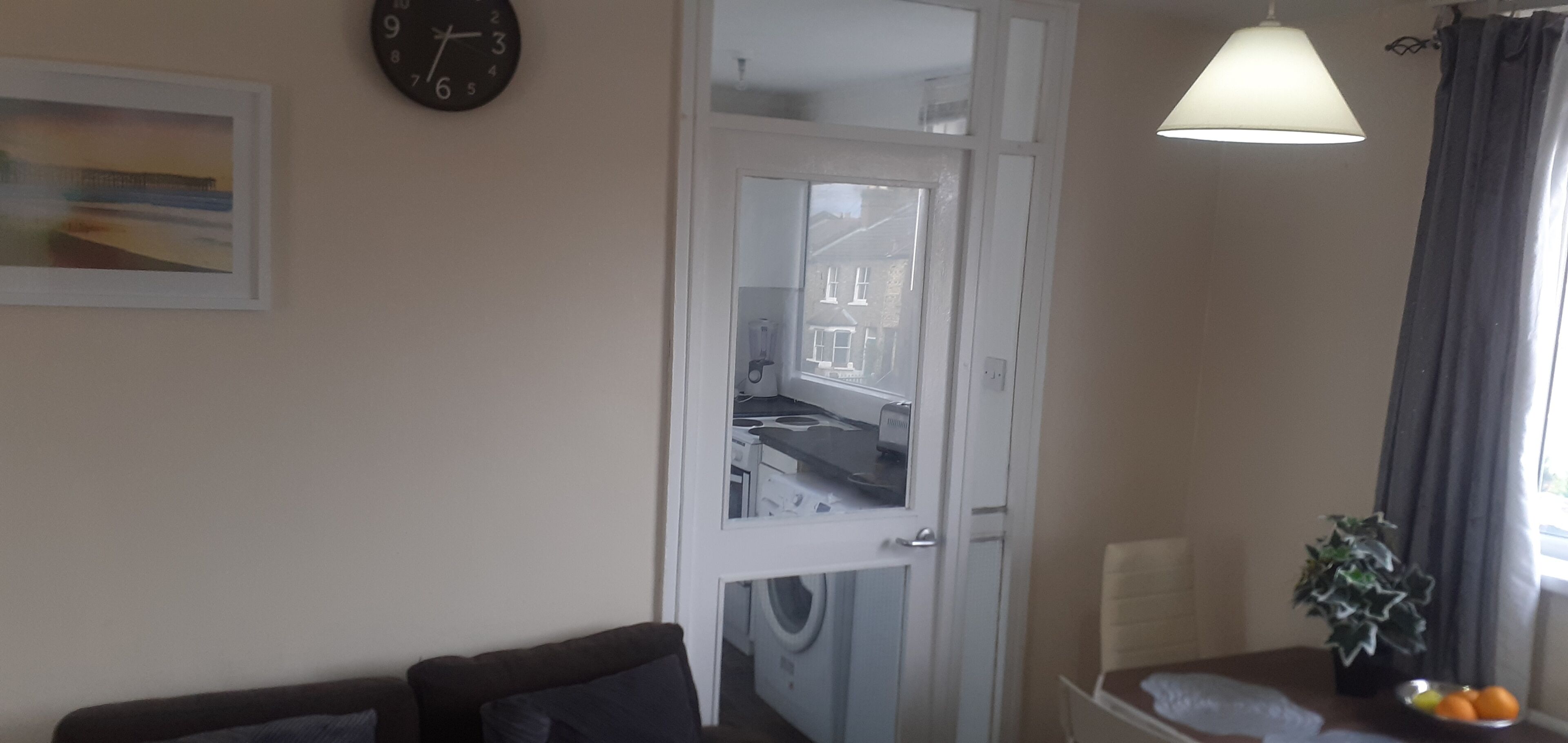 A room with a sofa, a clock, a door leading to a kitchen, a table with a plant and fruit, and a window with curtains.