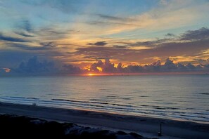 A beach scene with a sunset over the ocean, a clear sky, and a distant shoreline.