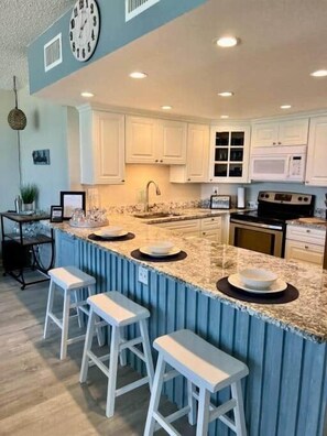 A kitchen with white cabinets, a granite countertop, a microwave, and a stove. There are three white bar stools.