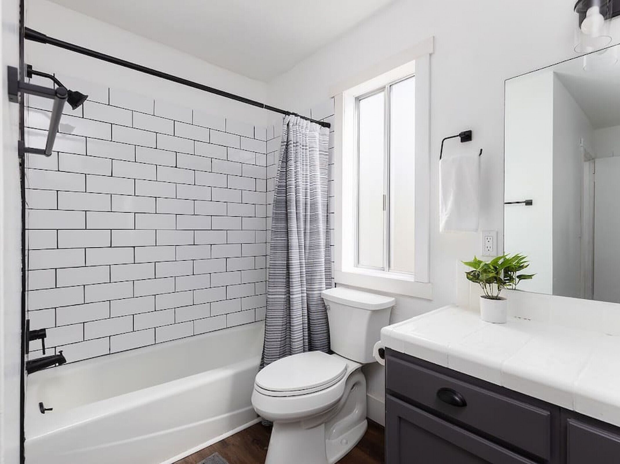 Une salle de bain moderne avec un mur en briques blanches, une douche avec un rideau à rayures, des toilettes blanches, un meuble-lavabo avec un miroir et une petite plante en pot.