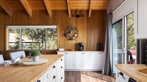A modern kitchen with wooden cabinets, a large window, and a dining area with a wooden table and chairs. - Whispering Pines | Amazing Views | Family Fun (Lake Arrowhead)