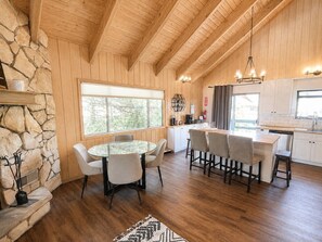 A wooden interior with a stone fireplace, a round dining table, bar stools, and a kitchen area.