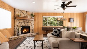 A cozy living room with a stone fireplace, a ceiling fan, and a large window. - Whispering Pines | Amazing Views | Family Fun (Lake Arrowhead)