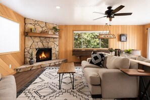 A cozy living room with a stone fireplace, a ceiling fan, and a large window.