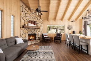 A cozy living room with a stone fireplace, wooden ceiling beams, a sofa, and a dining area with chairs and a table.