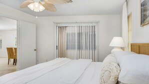 Ein Schlafzimmer mit Bett, Deckenventilator, Fenster mit durchsichtigen Vorhängen, Nachttisch mit Lampe und Korbstuhl.
