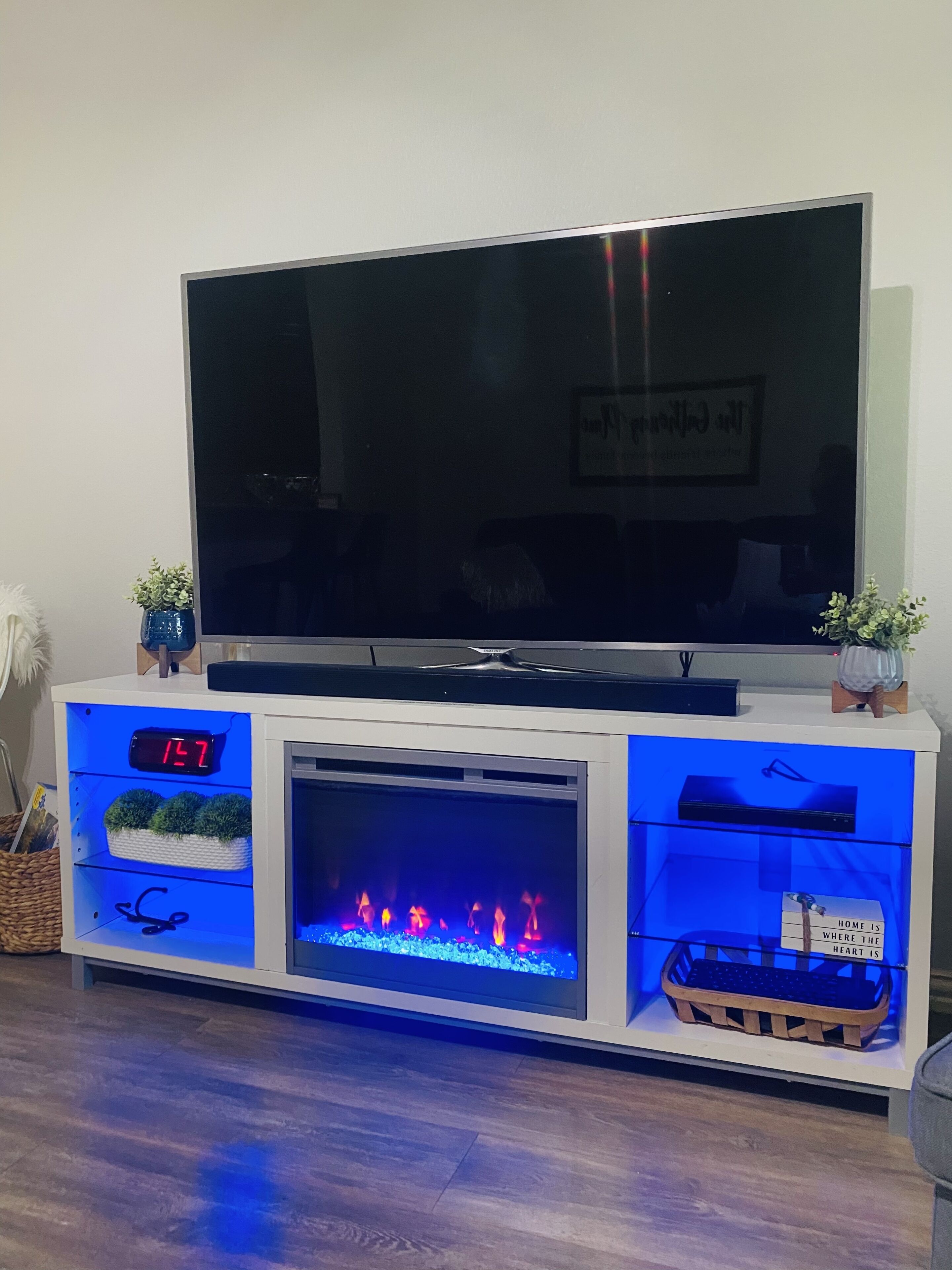 Un televisor de pantalla plana sobre un mueble blanco con chimenea empotrada, estantes y un reloj.