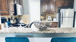 Une cuisine avec des meubles de rangement en bois, un four à micro-ondes, une cuisinière et un réfrigérateur. On y trouve deux tabourets de bar bleus et un bol décoratif en métal posé sur le comptoir.