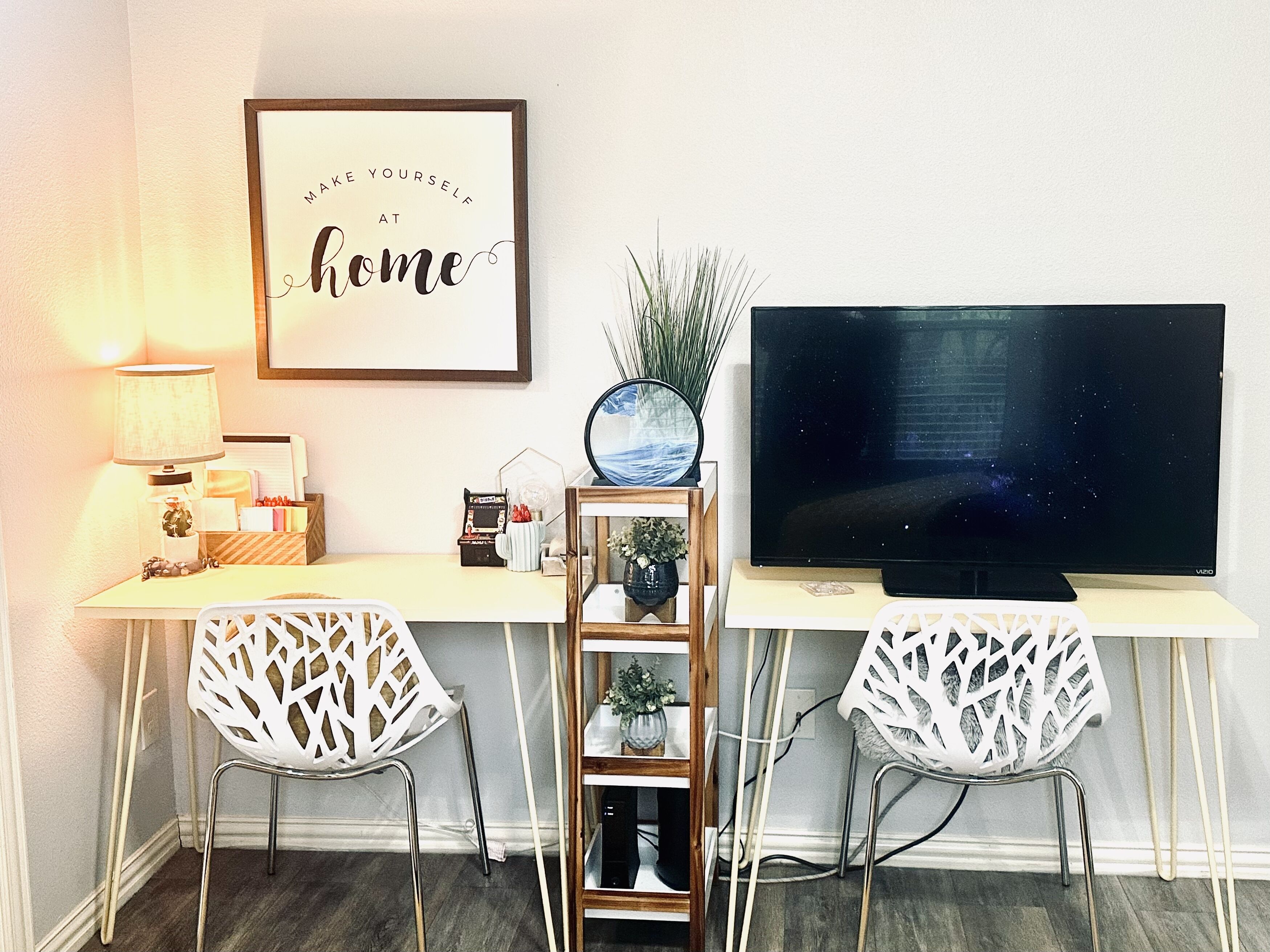 A home office setup with a desk, two white chairs, a TV, a framed inspirational quote, a lamp, and a small shelf with decorative items.