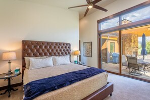 Une chambre à coucher avec une tête de lit capitonnée, un grand lit, une table de chevet avec une lampe, et une vue sur un patio extérieur avec une table et des chaises.