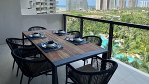 Un coin repas avec une table en bois dressée pour quatre personnes, des chaises noires et une vue sur une plage et des palmiers.