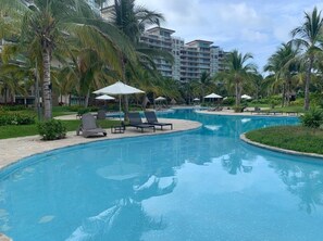 Ein Swimmingpool mit Liegestühlen und Sonnenschirmen, umgeben von Palmen und einem Hotel im Hintergrund.