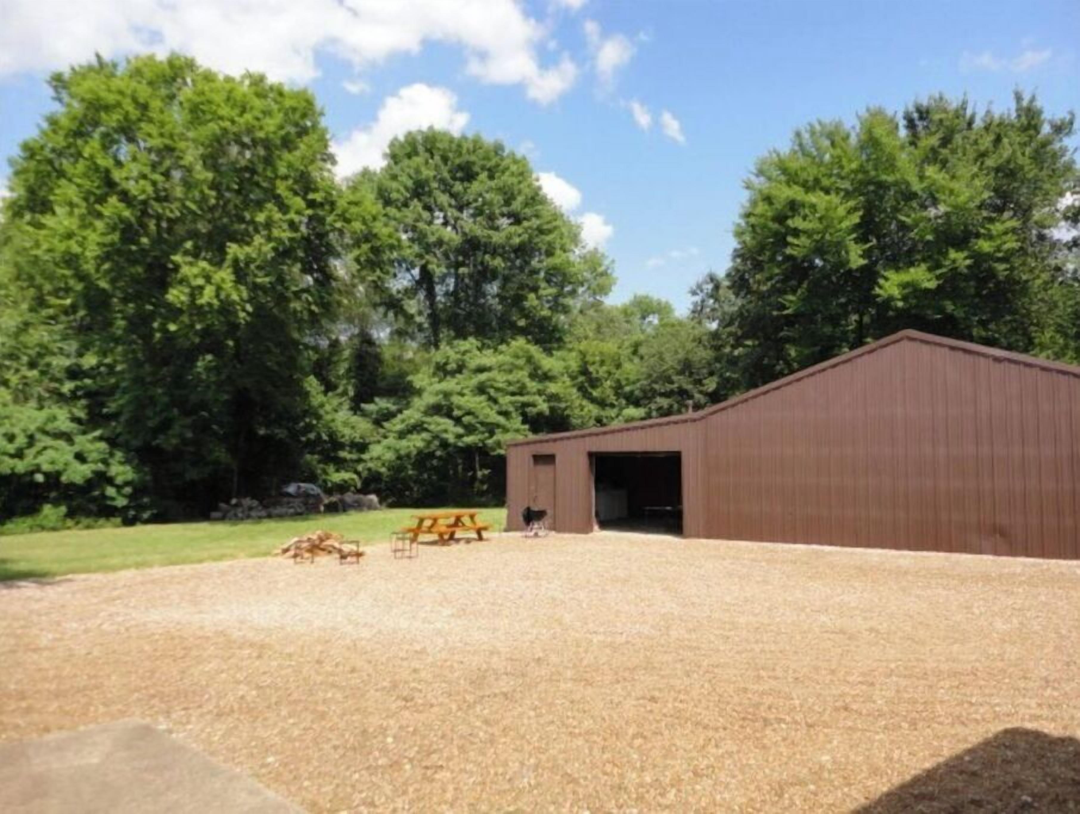 Un espace de stationnement recouvert de gravier, avec une table de pique-nique et un bâtiment brun, probablement une grange ou un abri de stockage, le tout entouré d’arbres.