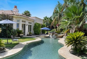 A swimming pool with a curved edge, surrounded by palm trees and a patio area with chairs and umbrellas.