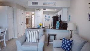 A compact living space with a kitchenette, two grey armchairs, a small table, and a sofa. - Hot Tub/Pool, Cov’d Parking (Gulf Shores)