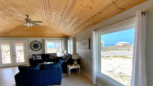 En rummelig stue med træloft, store vinduer med udsigt til stranden og en loftsvifte.