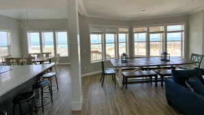 Un vaste espace de salle à manger avec vue sur la plage, une cuisine dotée d’un grand îlot central et un salon avec un canapé et des fauteuils.
