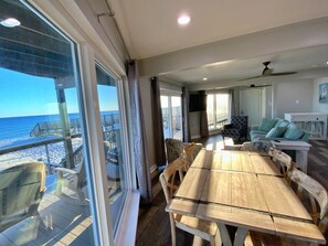 A spacious living area with a large wooden table and chairs, a sofa, and a television. A balcony with a view of the ocean is visible.