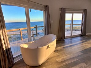 Une salle de bain spacieuse avec une grande baignoire, un plancher en bois et de grandes fenêtres offrant une vue sur la plage et l’océan.