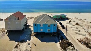 Ein Strandgrundstück mit drei Gebäuden, darunter ein großes blaues Haus mit Garage, ein kleineres grünes Haus und ein weißes Gebäude mit rotem Dach.