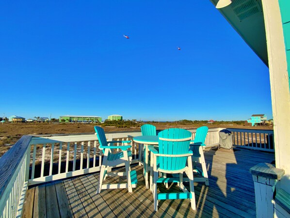 Une terrasse en bois, agrémentée de chaises Adirondack turquoise et d’une table, offre une vue sur une plage où l’on aperçoit des bâtiments au loin.