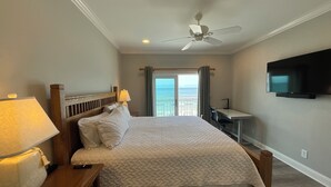 Ett hotellrum med en säng, ett skrivbord, en tv och utsikt över havet.