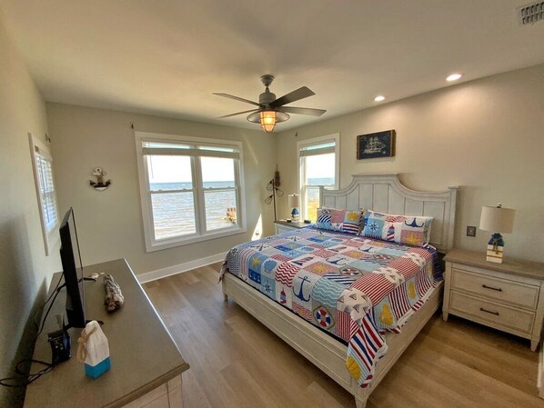 A bedroom with a bed, a desk, a chair, a ceiling fan, and a window with a view of the ocean.