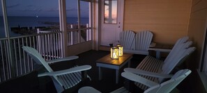 A balcony with white Adirondack chairs, a small table with a lantern, and a view of the ocean at dusk.