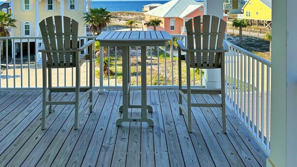 Une terrasse en bois avec deux fauteuils Adirondack et une petite table, donnant sur une plage bordée de maisons et sur l’océan.